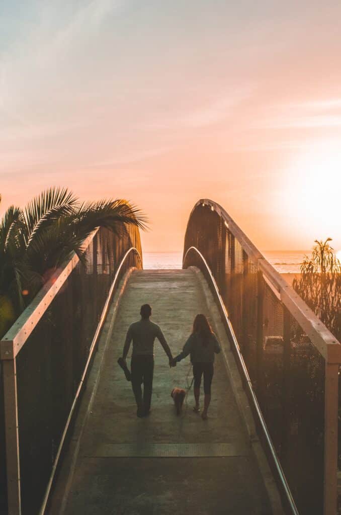 A man and a women walking a dog over a bridge with the sea and the sun in the end