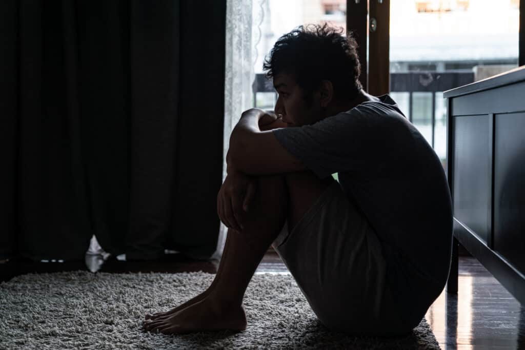 Depressed man struggling with drug addiction sitting on the floor with his knees to his chest