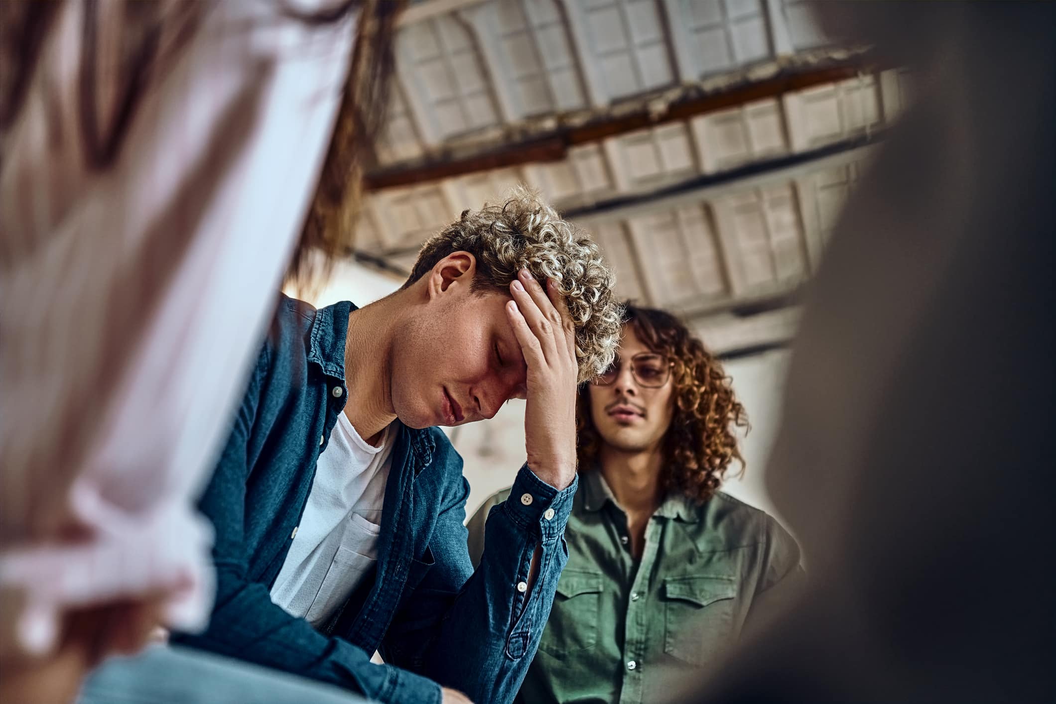 Close-up view of a devastated young man holding his head in his hands and a group of friends in a supportive pose around him.